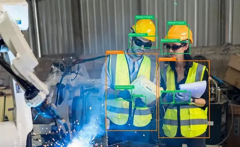 Two engineers in helmets and hi-vis vests beside a welding robot, each marked by PPE detection boxes