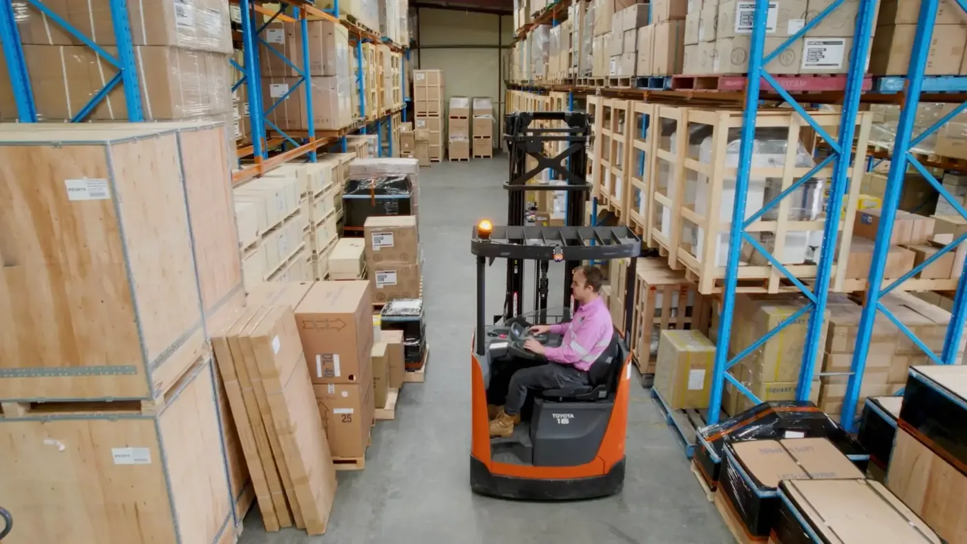 Operator driving an orange reach truck down a warehouse aisle lined with crates and racking