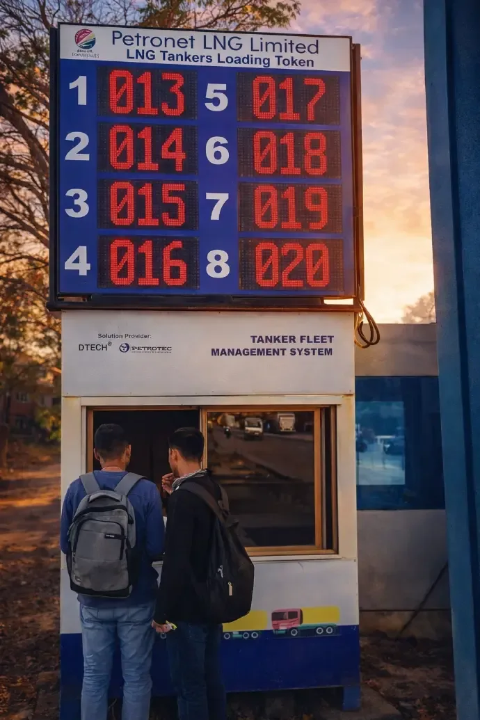 Two tanker drivers collecting tokens at the TFMS kiosk under the LED queue board